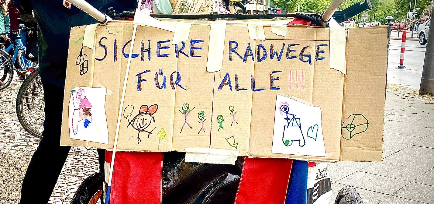Auf dem Foto ist ein Pappschild zu seinen, dass an der Rückseite eines Fahrradanhängers angebracht ist. Neben bunten Kinderzeichnungen ist in gemalten Großbuchstaben „sichere Radwege für alle“ zu lesen. 