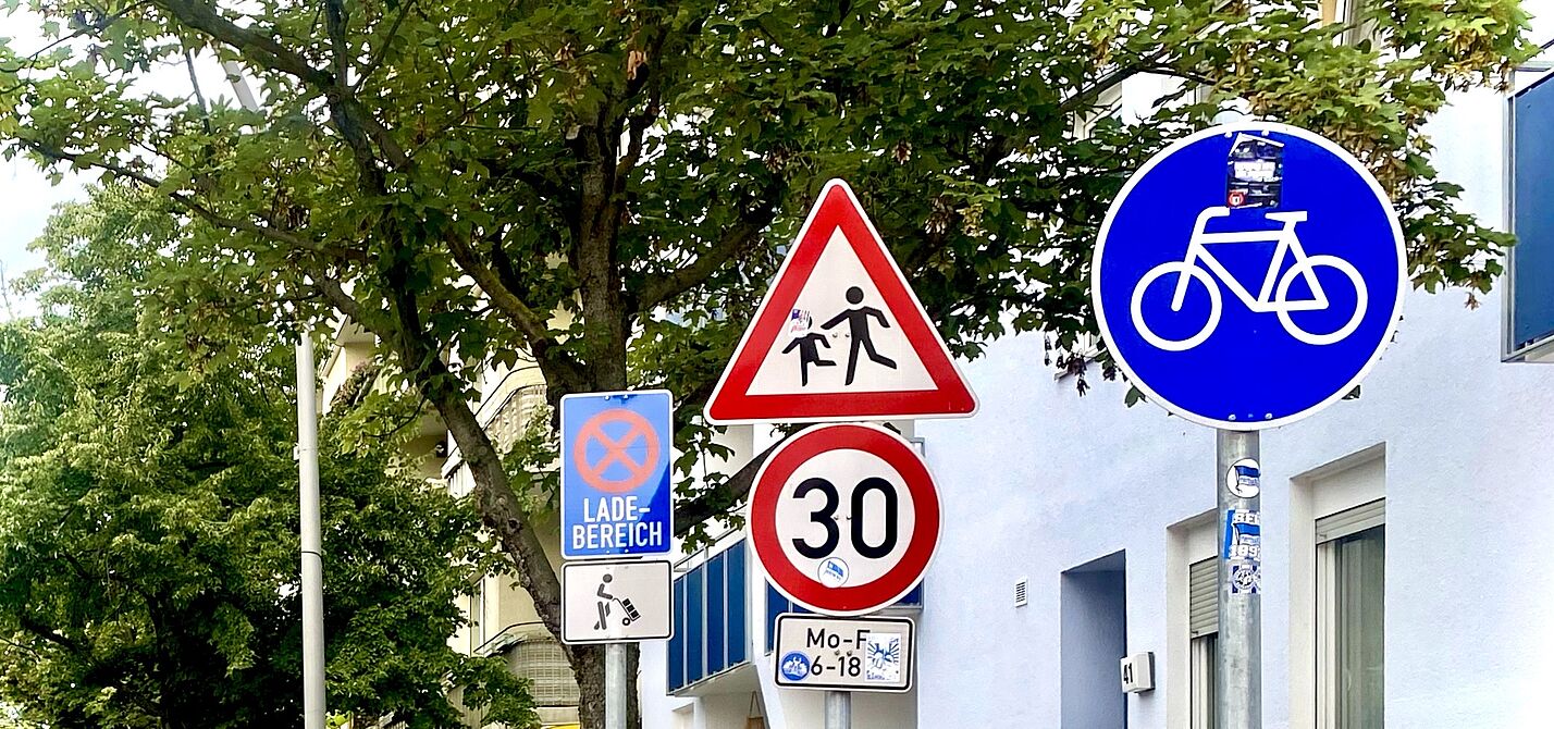 Auf dem Foto sind mehrere Verkehrszeichen zu sehen: Tempo 30, Kinder, benutzungspflichtiger Radweg, Lieferzone