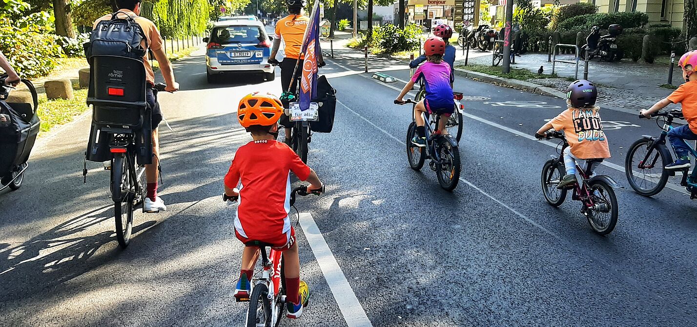 Kidical Mass Schöneberg September 2024 Eine Gruppe Kinder fährt bei sonnigem Wetter auf Fahrrädern das Berliner Reichpietschufer entlang, auf der Fahrbahn. Vorne fährt ein Auto der Polizei Berlin als Führungsfahrzeug, und zwei Erwachsene. Es ist eine ADFC Berlin Fahne zu sehen.