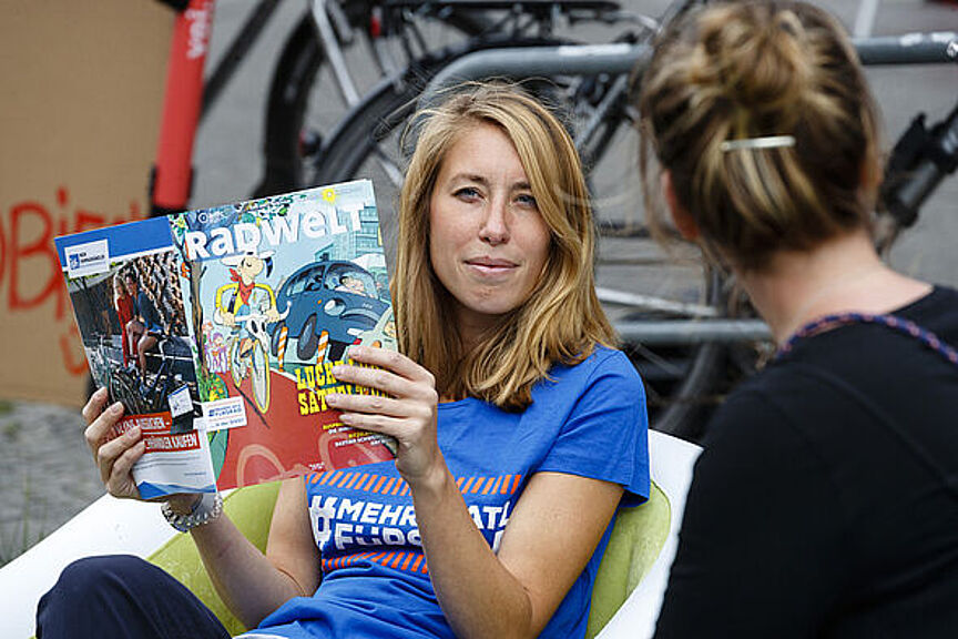 Willkommen im Frauen*Netzwerk des ADFC Berlin Eine Frau in einem ADFC T-Shirt sitzt auf einem Stuhl und liest die "radwelt"