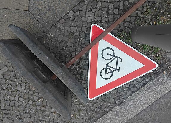 Radwegestopp 2.0? Das Bild zeigt ein rotes dreieckiges "Achtung Radfahrer"-Schild, es liegt auf dem Boden neben dem Fuß eines Baustellen-Aufstellers