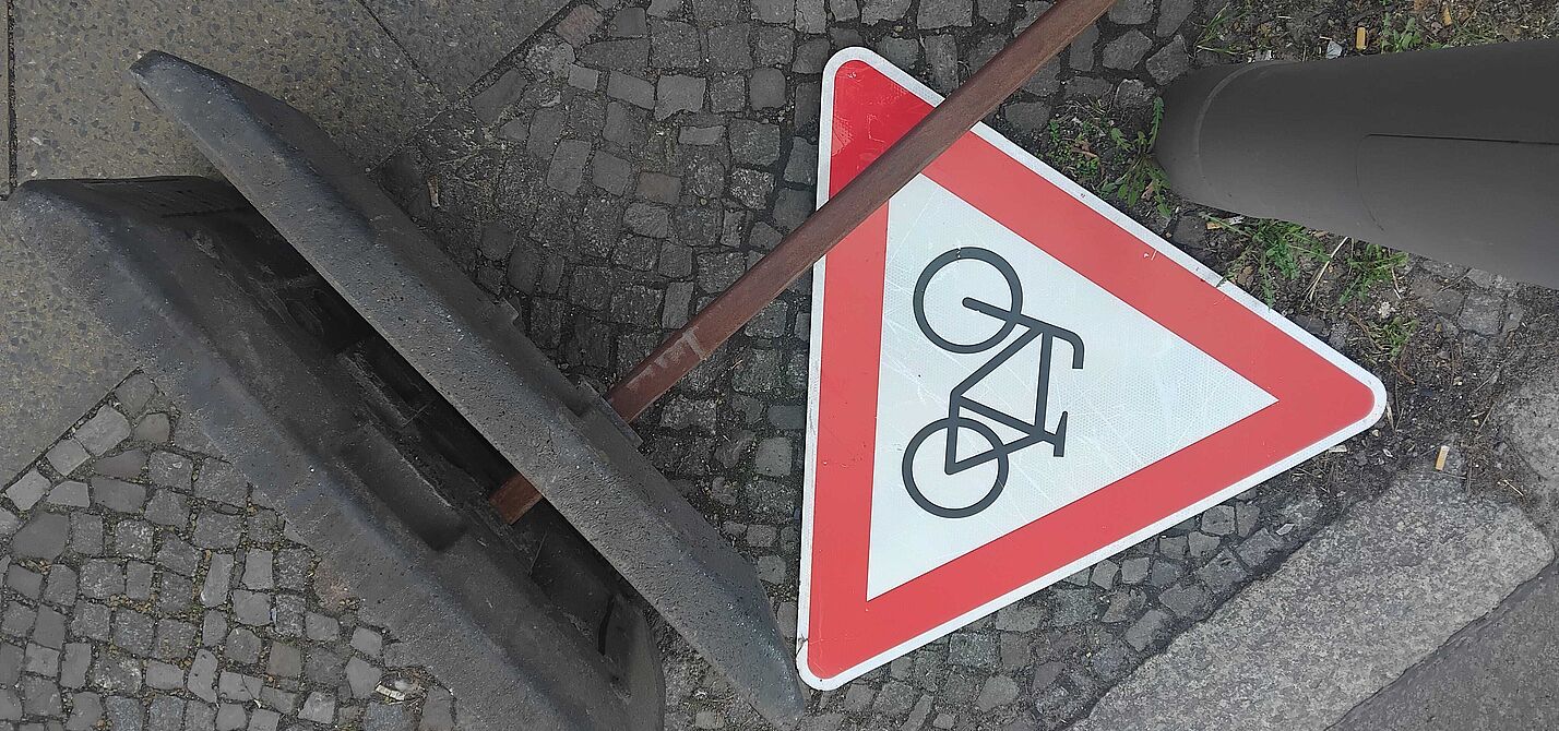 Das Bild zeigt ein rotes dreieckiges "Achtung Radfahrer"-Schild, es liegt auf dem Boden neben dem Fuß eines Baustellen-Aufstellers