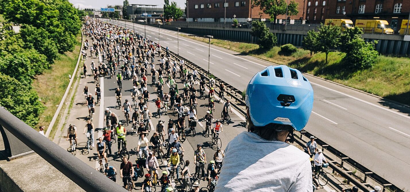 Kind mit blauem Helm guckt auf Autobahn über die auf einer Seite hunderte FahrradfahrerInnen fahren