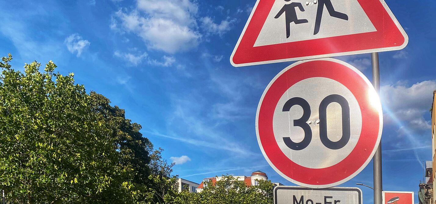 Tempo 30 auf dem Schulweg an einer Hauptverkehrsstraße: Grunewaldstraße in Berlin Schöneberg Auf dem Foto sind zwei Verkehrsschilder vor blauem Himmel zu sehen: Tempo 30 und Kinder. Rechts liegt eine Häuserzeile, auf der linken Seite ist die Straße durch Bäume begrünt.