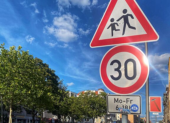 Tempo 30 auf dem Schulweg an einer Hauptverkehrsstraße: Grunewaldstraße in Berlin Schöneberg Auf dem Foto sind zwei Verkehrsschilder vor blauem Himmel zu sehen: Tempo 30 und Kinder. Rechts liegt eine Häuserzeile, auf der linken Seite ist die Straße durch Bäume begrünt.