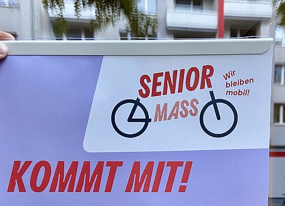 Ein Plakat mit dem Titel „Senior Mass – Kommt mit!“ wird vor einer Schaufensterscheibe ins Licht gehalten.