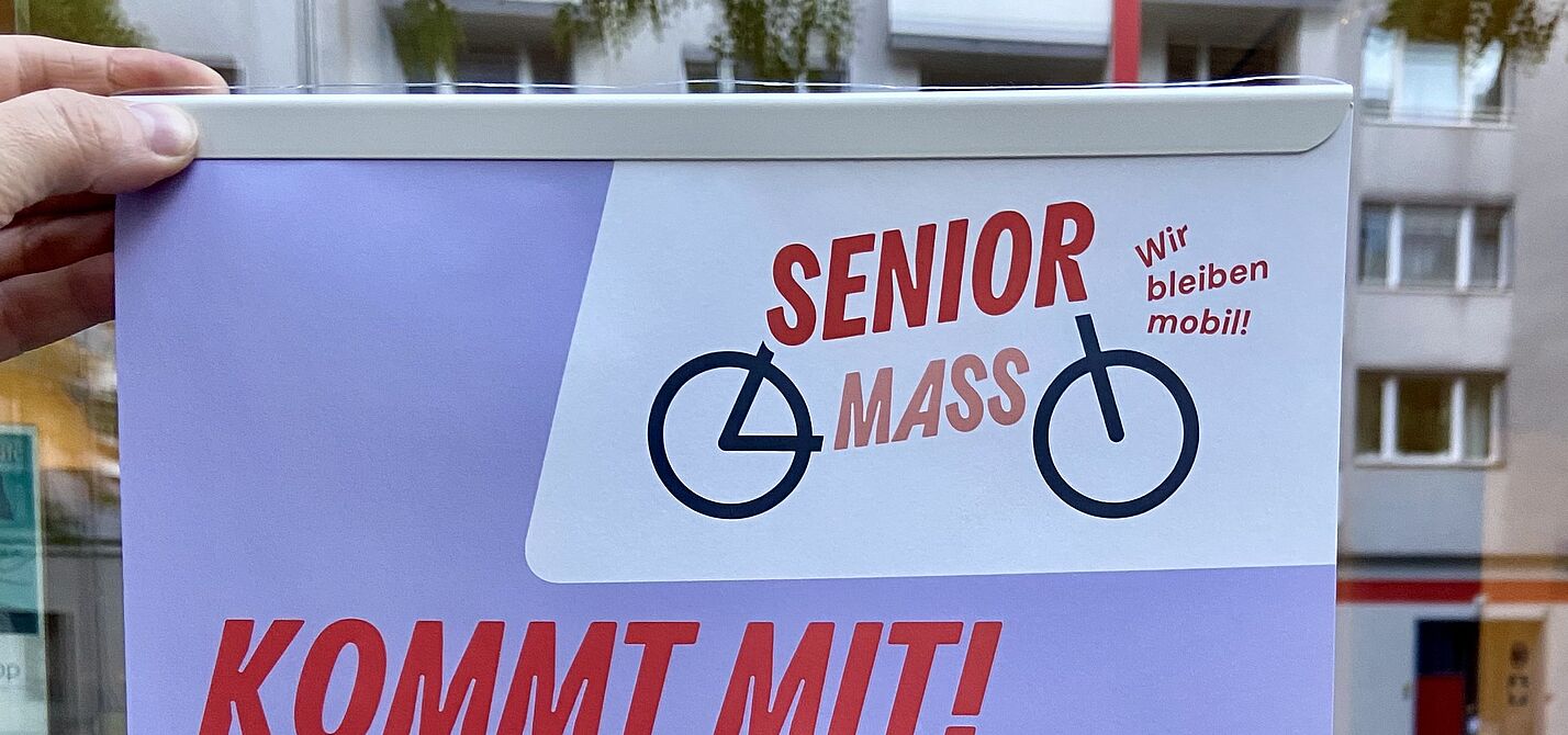 Ein Plakat mit dem Titel „Senior Mass – Kommt mit!“ wird vor einer Schaufensterscheibe ins Licht gehalten.