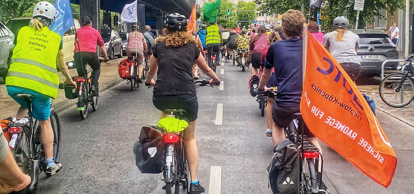 Eine Gruppe Radfahrende fährt mit Fahnen auf einer Fahrbahn Richtung Kreuzberg. Links im Bild ist die oberirdische U-Bahnlinie 1 zu sehen. 