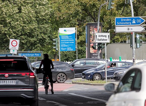 Auf einer Hauptverkehrsstraße am Heidelberger Platz in Berlin ist unter einer Brücke nahe einer Auffahrt zur A100 die Silhoutte eines Radfahrers zu sehen. Er steht mit seinem Rad mitten auf der Fahrbahn, umgeben von Autos. Zum Radfahren ist dort wenig Platz. Es sind mehrere Schilder zu sehen, unter anderem ein weißes Schild, das die Aufschrift „Achtung Radfahrer“ trägt.