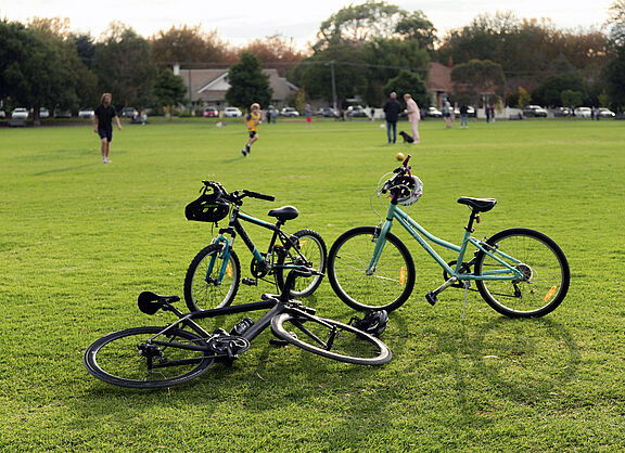 Im Vordergrund sind drei Räder stehend und liegend auf einer Rasenfläche zu sehen, im Hintergrund sind ein paar Menschen auf dem Rasen stehend mit einem Fußball zu sehen.