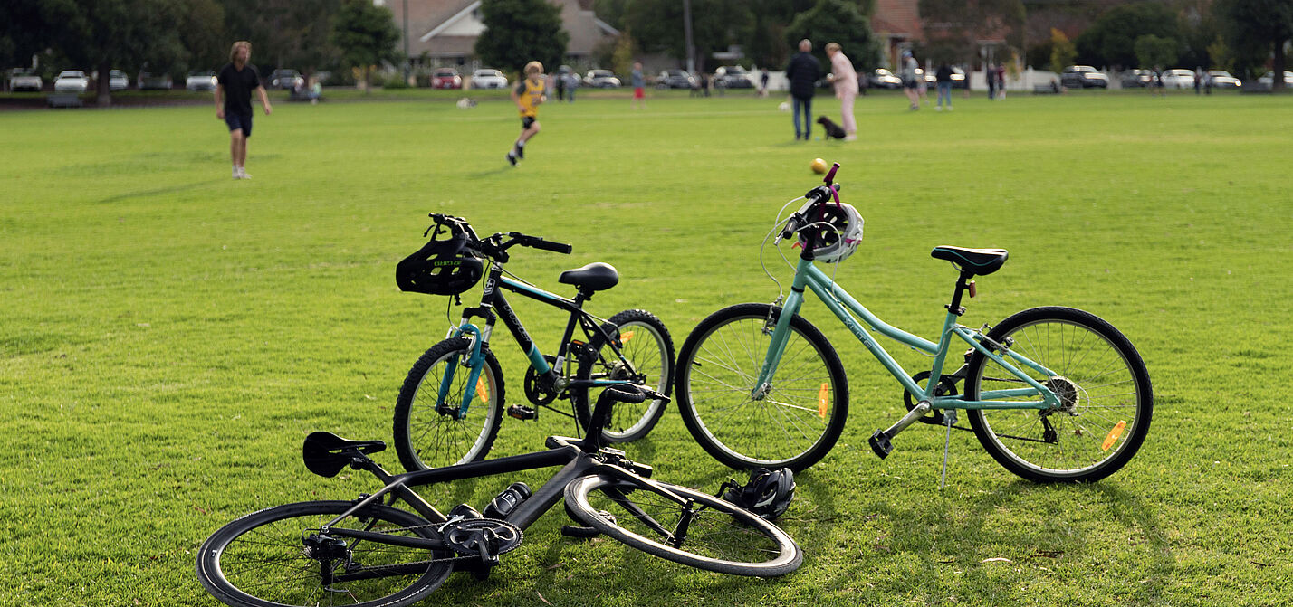 Im Vordergrund sind drei Räder stehend und liegend auf einer Rasenfläche zu sehen, im Hintergrund sind ein paar Menschen auf dem Rasen stehend mit einem Fußball zu sehen.