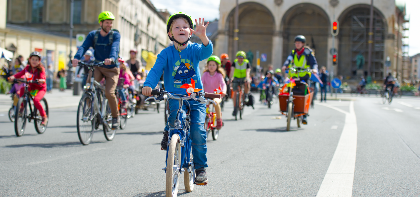 Kinder fahren mit dem Fahrrad bei einer Familien-Fahrraddemo. Ein Kind im Vordergrund winkt in die Kamera.